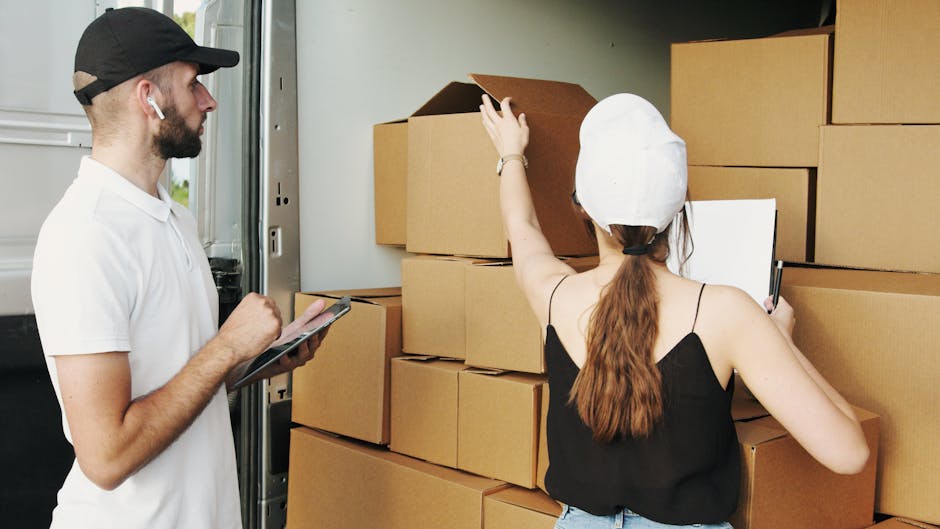 A man wearing a black cap and white polo shirt is kneeling on the pavement next to a partially packed cardboard moving box, inside which he is writing with a black marker. Surrounding him are several