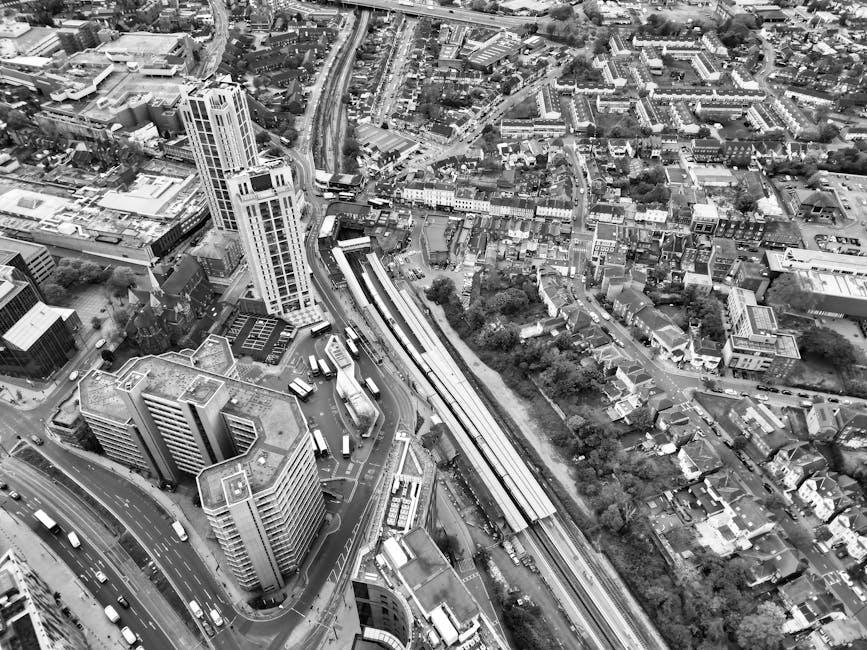 Aerial black-and-white photograph showing a dense urban area with a mix of high-rise office buildings, residential structures, and smaller houses. Prominent in the image is a railway station with platforms and tracks running parallel to a curved road, with multiple buses and cars visible on the streets. Surrounding the station are tall office buildings with flat roofs, some featuring rooftop equipment, and smaller commercial and residential buildings. The terrain is relatively flat, with a network of roads and pathways connecting the area. Trees and green spaces are scattered among the buildings. The photograph captures a busy cityscape with developed infrastructure, typical of a central business district, emphasizing the proximity of transport links and urban density. This image represents a typical urban environment that remains under the framework of city planning and transport logistics, relevant to house removals and furniture transport in the Dagenham area, as seen in the context of [PAGE_TITLE] by [COMPANY_NAME].