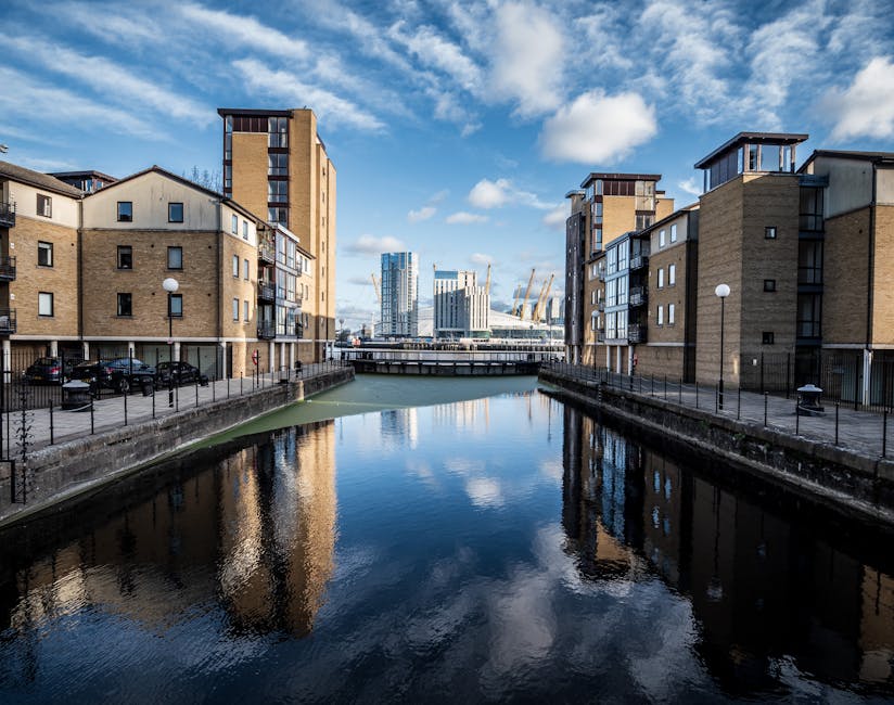 A daytime view of a canal flanked by modern, multi-storey residential buildings with beige brick facades, large windows, and small balconies. A narrow paved pathway runs alongside the canal, lined with black metal railings and evenly spaced street lamps. In the foreground, the calm water of the canal reflects the buildings and the partly cloudy sky above. In the distance, a bridge crosses over the canal, and further ahead, a cluster of tall, contemporary office buildings with glass exteriors and construction cranes can be seen, indicating ongoing development in the area. This urban scene illustrates a well-maintained residential and commercial waterfront environment, suitable for home relocation and furniture transport services such as those offered by Man and Van Dagenham, which often assist with packing and loading during house removals.