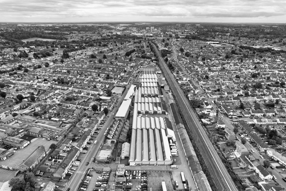 Aerial black-and-white photograph showing a dense urban area with a mix of high-rise office buildings, residential structures, and smaller houses. Prominent in the image is a railway station with platforms and tracks running parallel to a curved road, with multiple buses and cars visible on the streets. Surrounding the station are tall office buildings with flat roofs, some featuring rooftop equipment, and smaller commercial and residential buildings. The terrain is relatively flat, with a network of roads and pathways connecting the area. Trees and green spaces are scattered among the buildings. The photograph captures a busy cityscape with developed infrastructure, typical of a central business district, emphasizing the proximity of transport links and urban density. This image represents a typical urban environment that remains under the framework of city planning and transport logistics, relevant to house removals and furniture transport in the Dagenham area, as seen in the context of [PAGE_TITLE] by [COMPANY_NAME].