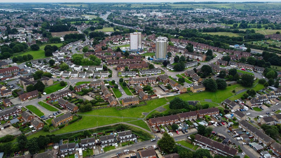 An aerial view of a residential neighbourhood in Becontree estate, Dagenham, showing rows of terraced houses, semi-detached homes, and apartment buildings surrounded by green spaces, parks, and tree-lined streets. The image captures a driveway with parked cars and a recent home relocation scene where a man and a woman, dressed casually, are loading large cardboard boxes and wrapped furniture into a blue moving van parked on the street. The van is positioned at the curb, with its rear doors open, revealing packing and moving equipment such as blankets, straps, and trolleys. The surrounding area includes pathways, small lawns, and a mixture of low-rise buildings and taller apartment blocks, reflecting the typical urban residential setting involved in furniture transport and house removals within Dagenham, as seen on the manandvandagenham.org.uk removals services page.