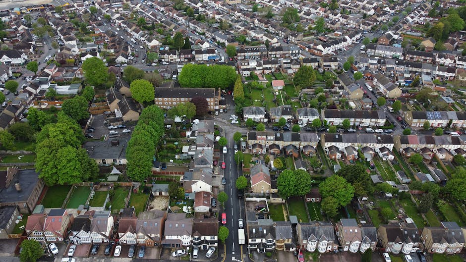 Aerial view of a residential neighbourhood in Becontree estate, Dagenham, showing a dense layout of terraced and semi-detached houses with rooftops, gardens, and tree-lined streets. Parked cars are visible along the narrow streets, while larger vehicles and moving boxes are seen near the entrances of some homes, indicating a home relocation process. The scene includes a variety of building materials such as tiled roofs, brick walls, and wooden fences, with some properties featuring small front yards and driveways. The overall environment is well-lit with natural daylight, highlighting the orderly arrangement of houses and the presence of green spaces. The image conveys the context of house removals and the logistics involved in furniture transport and packing within a typical suburban setting, as curated by Man and Van Dagenham for the Becontree estate moves.