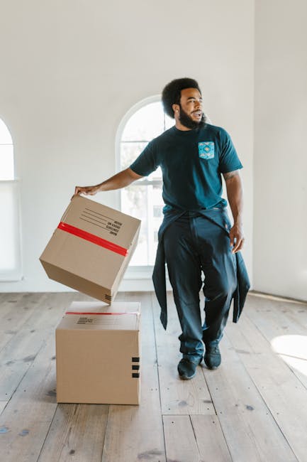 A man with dark curly hair and a beard is inside a well-lit room with white walls and a large arched window, carrying a cardboard box wrapped with red tape in his right hand. He is dressed in a navy blue t-shirt featuring a blue logo on the chest and dark trousers, with a jacket tied around his waist. Behind him, on the light wooden floor, there are two additional cardboard boxes, one stacked on top of the other, ready for loading or unpacking during a house relocation. The scene captures the process of packing and moving, highlighting the careful handling of boxed items as part of furniture transport and home moving services. The room appears empty aside from the boxes, suggesting it is in the midst of a removal or delivery operation. The overall setting emphasizes professionalism in domestic moving activities, as seen with [COMPANY_NAME], operating in Dagenham, and aligns with the service of house removals described in the page titled 'Dagenham removals real cost and what to expect'.