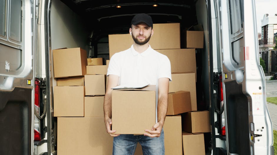 Two individuals, a man and a woman, are engaged in a home relocation process involving a white moving van with its rear doors open. The man, dressed in a white t-shirt, blue jeans, and a dark baseball