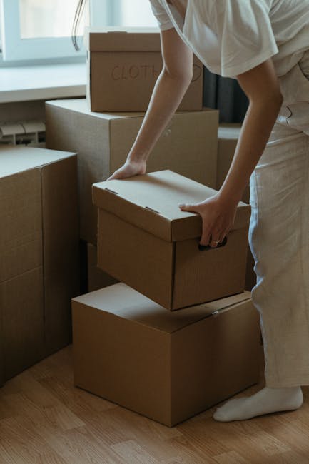 A person wearing light-colored clothing is in the process of packing or unpacking a home during a house move, shown stacking and carrying plain cardboard storage boxes inside a well-lit room near a wi