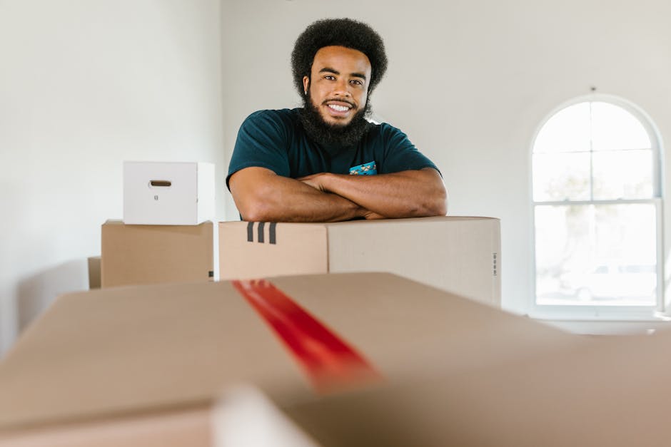 A person wearing light-colored clothing is in the process of packing or unpacking a home during a house move, shown stacking and carrying plain cardboard storage boxes inside a well-lit room near a wi